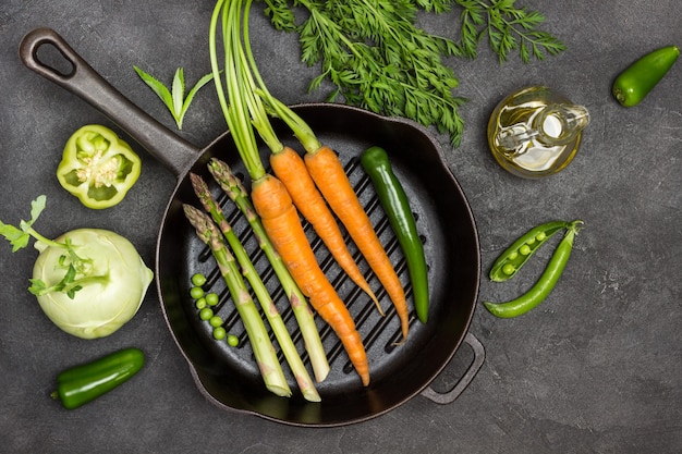 Carottes fraîches avec fanes vertes, gousse de poivron vert et asperges dans une poêle à frire. Bouteille d'huile d'olive, chou-rave, poivrons et pois verts sur table. Fond noir. Mise à plat