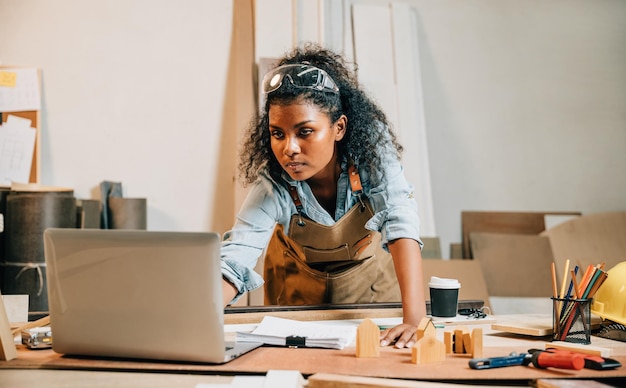 Carpenter America femme noire aux cheveux bouclés croquis prenant des notes dans un papier de travail tout en se tenant à une table en bois avec un ordinateur portable jeune femme travaillant et apprenant en ligne dans un atelier de menuiserie Happy Carpenters Day