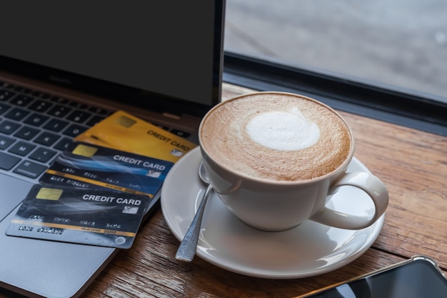 Carte de crédit d'un ordinateur portable, d'un smartphone et d'un café au lait chaud avec de la mousse de lait d'art latte dans une tasse sur fond de bois, concept de banque en ligne