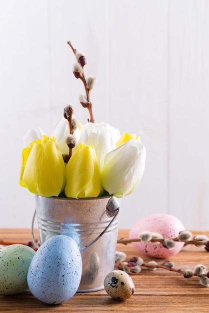 Carte de Joyeuses Pâques avec des fleurs de tulipes fraîches au printemps dans un seau et des œufs peints à la main sur une table en bois.