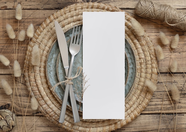 Carte de menu vierge sur assiette avec fourchette et couteau sur table en bois avec décorations bohèmes et plantes séchées, vue de dessus. Maquette de carte de mariage Boho