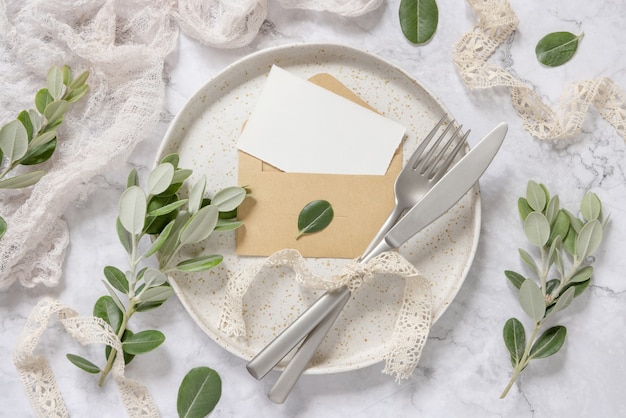 Carte de papier vierge dans une enveloppe posée sur une plaque blanche avec une fourchette et un couteau sur une table en marbre avec des branches d'eucalyptus et des rubans vintage autour, vue de dessus. Maquette de carte