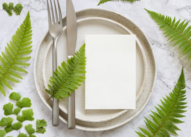 Carte de papier vierge posée sur une assiette avec fourchette et couteau sur une table en marbre avec des feuilles de fougère autour de la vue de dessus. Scène de maquette tropicale avec carte d'invitation à plat