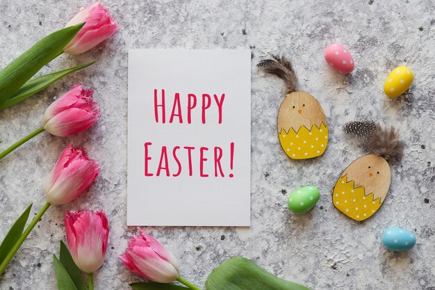 Carte de Pâques avec tulipes roses jouets de poulet en bois oeufs colorés et papier avec texte JOYEUSES PÂQUES