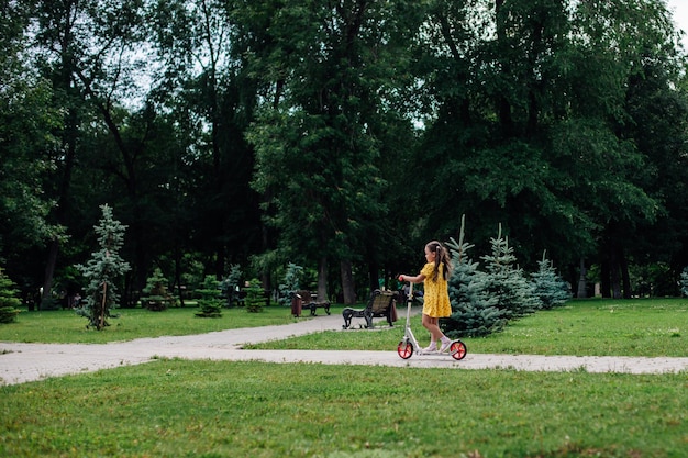 Carte postale d'été une fille fait du scooter dans un parc d'été verdoyant avec des arbres et une pelouse verte sur le bac...