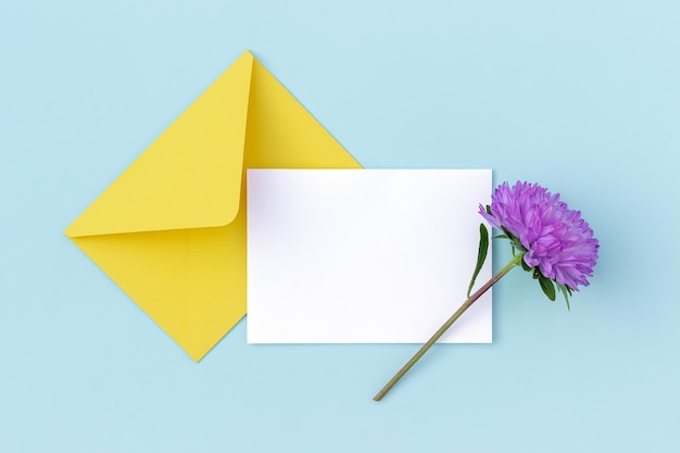 Photo carte vierge blanche enveloppe jaune et fleur violette sur fond bleu style minimal vue de dessus mise à plat maquette