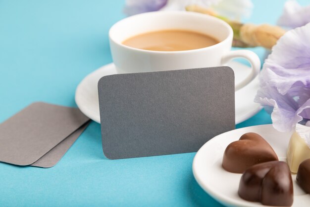 carte de visite grise avec tasse de cioffee, bonbons au chocolat et fleurs d'iris sur fond bleu pastel