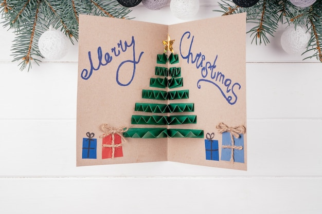 Carte de voeux en carton fait main avec arbre de Noël 3D en accordéon plié en papier coloré, avec inscription Joyeux Noël sur fond blanc avec des branches de sapin. Vue de dessus