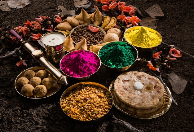 Carte de voeux Happy Holi conçue montrant des aliments traditionnels indiens sucrés et salés, des fleurs et des couleurs en poudre disposées sur fond blanc ou argile. Mise au point sélective