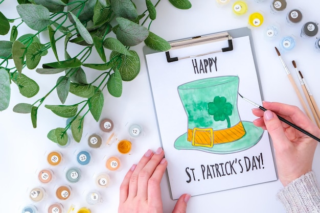 Carte de voeux peinte maison St Patricks day. Idée cadeau, déco. DIY. Fais le toi-même. Trèfles, pinceau et peinture. Mains féminines. Chapeau de la Saint Patrick.