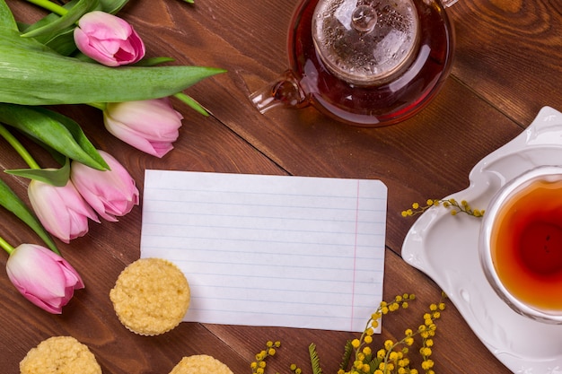 Carte de voeux pour le jour féminin avec tulipes, mimosa, thé et cupcakes sur fond en bois marron.