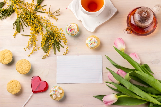 Carte de voeux de printemps avec tulipes, mimosa, thé et cupcakes sur table en bois.