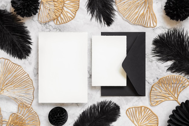 Cartes et enveloppe sur une table en marbre près de plumes noires et de feuilles dorées Maquette de mariage