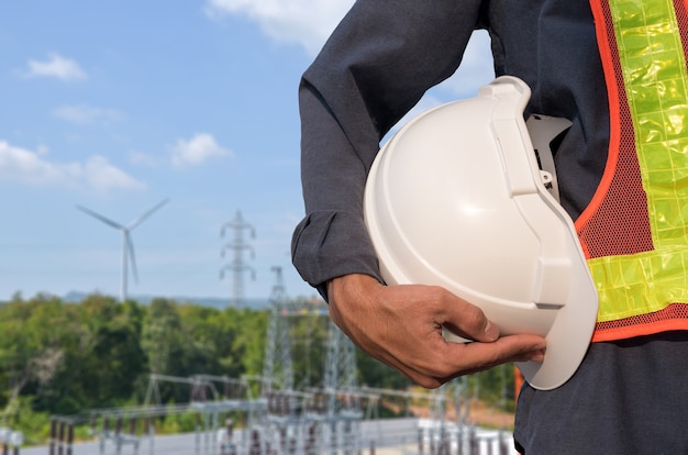 Le casque blanc est entre les mains d'un électricien sur fond de centrale éolienne.