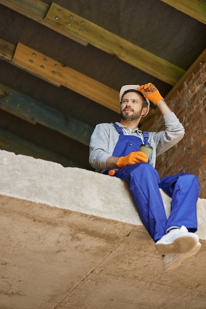 Casque de chantier jeune homme positif à l'air joyeux, prenant une pause et buvant du café à l'intérieur