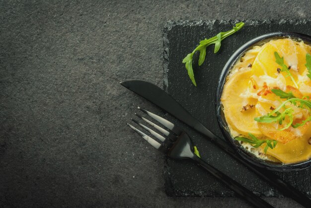 Casserole. Cuisine française. Gratin de pommes de terre fait maison dans un bol pour la cuisson. Sur un fond de pierre noire. Avec des feuilles de roquette fraîche pour la décoration. Copyspace vue de dessus