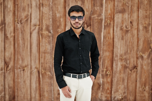 Casual jeune homme indien en chemise noire et lunettes de soleil posés sur fond de bois.