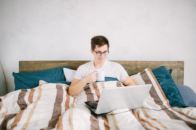 Casual jeune homme utilisant un ordinateur portable dans son lit à la maison et buvant du café