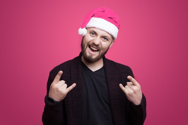 Casuca barbu fait des gestes le signe du rock and roll en souriant sur un mur rose portant un chapeau de père Noël