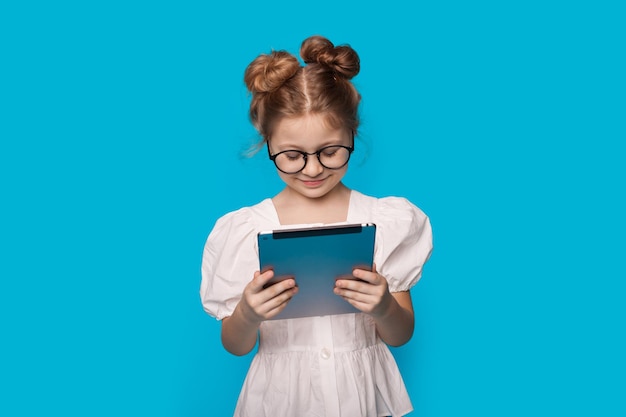 Caucasian cute girl lit à partir d'une tablette en souriant sur un mur de studio bleu à travers des lunettes dans une robe blanche