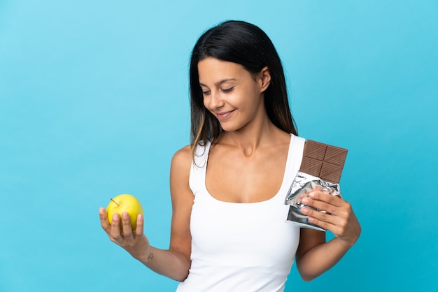 Caucasian girl isolated on blue prenant une tablette de chocolat dans une main et une pomme dans l'autre