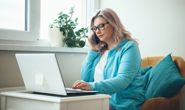 Caucasian senior businesswoman avec des lunettes et des cheveux blonds parle au téléphone tout en travaillant à distance avec un ordinateur portable