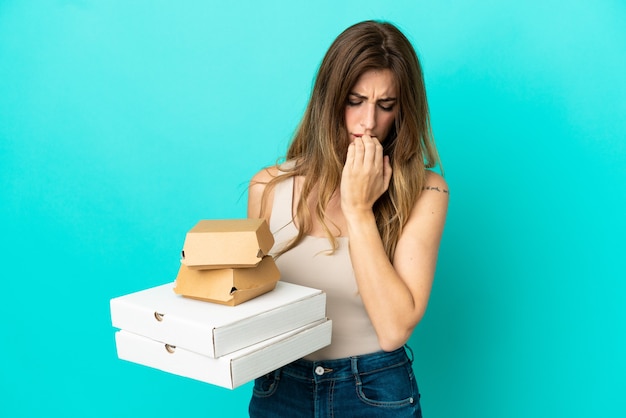 Caucasian woman holding pizzas et burger isolé sur fond bleu ayant des doutes