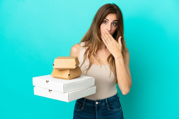 Caucasian woman holding pizzas et burger isolé sur fond bleu heureux et souriant couvrant la bouche avec la main