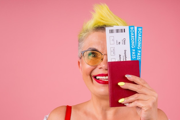 Photo caucasien belle vieille senior avec des cheveux teints en jaune élégants et des lunettes de soleil tenant un passeport et un billet d'avion sur fond de studio rose. lèvres rouges pour toujours jeune âme.