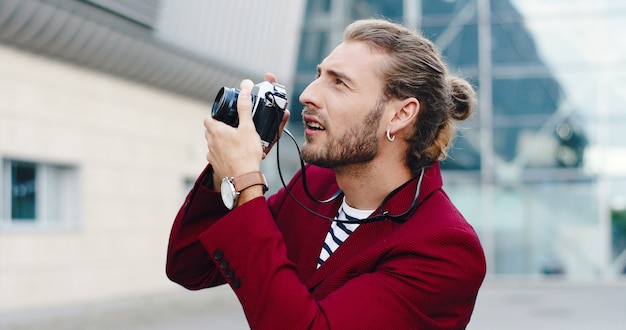 Caucasien jeune beau photographe masculin élégant prenant des photos avec un appareil photo en plein air dans la ville d'affaires. Sourire homme gai faisant des photos de l'architecture urbaine de la ville.