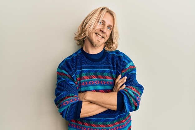 Caucasien jeune homme aux cheveux longs portant un chandail d'hiver coloré visage heureux souriant avec les bras croisés en regardant la caméra. Personne positive.