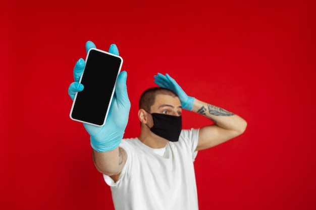 Caucasien jeune homme en masque protecteur et gants médicaux sur le mur du studio rouge montre l'écran du téléphone vide