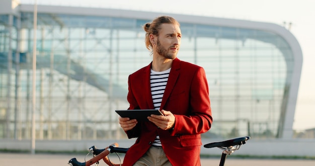 Caucasien jeune a souri bel homme debout à vélo et à l'aide de tablette. Gars élégant en veste rouge s'appuyant sur le vélo et tapotant ou faisant défiler sur un ordinateur gadget au paysage de la ville à l'aéroport.