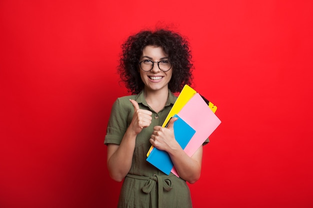 Caucasienne femme aux cheveux bouclés tient des livres et fait des gestes le signe similaire tout en posant sur un mur rouge