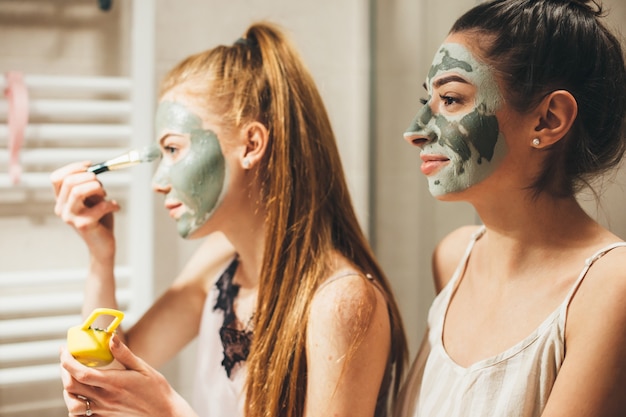 Caucasienne femme aux cheveux roux et son amie brune appliquent un masque anti-acné du visage tout en regardant dans le miroir