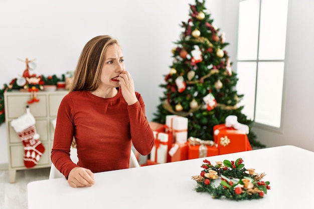 Caucasienne jeune femme blonde assise sur la table près de l'arbre de noël à l'air stressée et nerveuse avec les mains sur la bouche se ronger les ongles. problème d'anxiété.