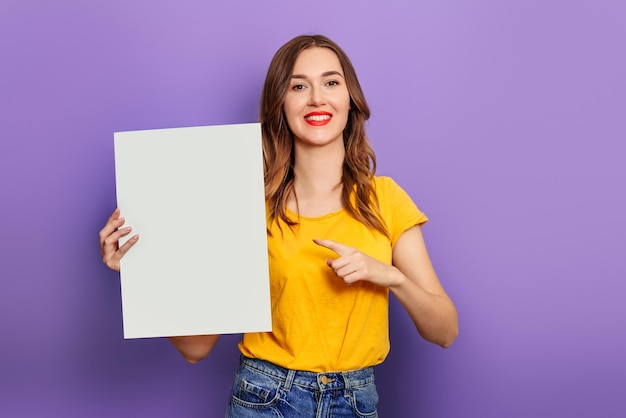 Caucasienne jeune femme tenant une affiche blanche dans ses mains portant un t-shirt jaune et isolée sur un fond lilas. maquette