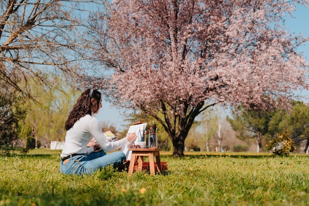 Caucasienne jeune fille aux cheveux ondulés peignant une toile avec des peintures rose pastel en se tenant debout dans la nature d'un parc, devant un amandier rose géant.