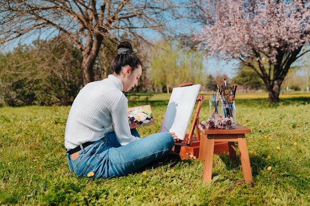 Caucasienne jeune fille aux cheveux ondulés, peindre une toile avec des peintures rose pastel en se tenant debout dans la nature d'un parc