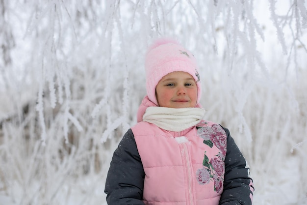 Caucasienne jeune fille aux joues vermeilles rouges dans des vêtements d'hiver chauds regardant la caméra légèrement souriante avec des arbres enneigés en arrière-plan Promenade d'hiver en forêt