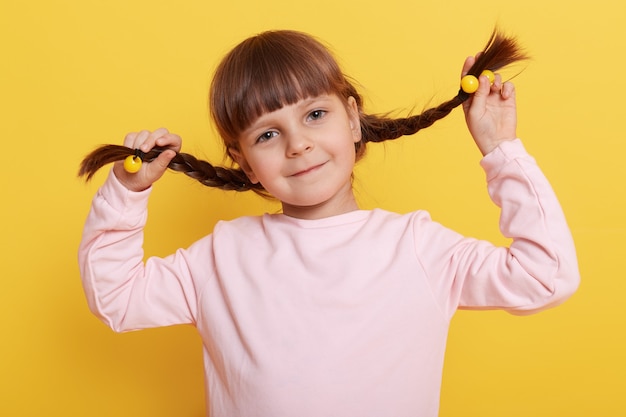 Caucasienne petite fille se tirant en nattes tout en regardant directement avec joli sourire charmant, enfant en chemise rose pâle décontractée isolé sur mur jaune.