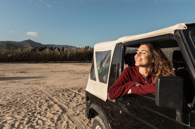 Caucasienne souriante jeune femme aux yeux fermés regardant par la fenêtre. Voyage en véhicule tout-terrain 4x4. Fond de désert de sable et de plage.