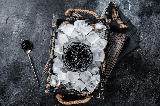 Caviar noir servi avec de la glace dans un bol en verre, cuillère à caviar noir. Fond noir. Vue de dessus.