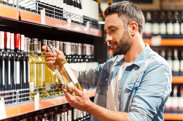 Cela devrait être bien. Vue latérale d'un beau jeune homme tenant une bouteille de vin et la regardant en se tenant debout dans un magasin de vin