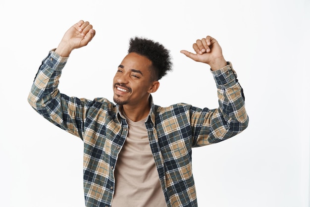 Célébration Heureux homme afro-américain dans des vêtements décontractés se sentant libre et joyeux à la fête levant les mains en souriant avec les yeux fermés dansant debout sur fond blanc