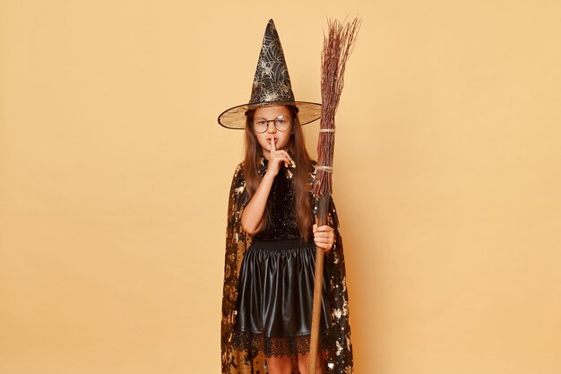 Photo célébration mystique carnaval magique assistant énigmatique sorcière effrayante petite fille en manteau noir avec balai isolé sur fond beige garde le secret avec un visage mystérieux