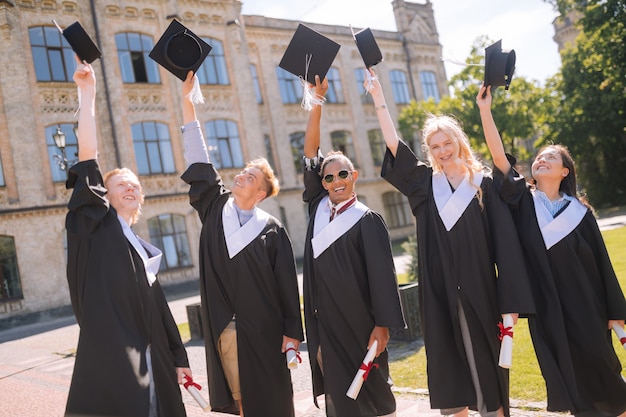 Célébration de la remise des diplômes. De joyeux diplômés debout dans la cour de l'université et levant les mains avec des casquettes de maître en l'air.