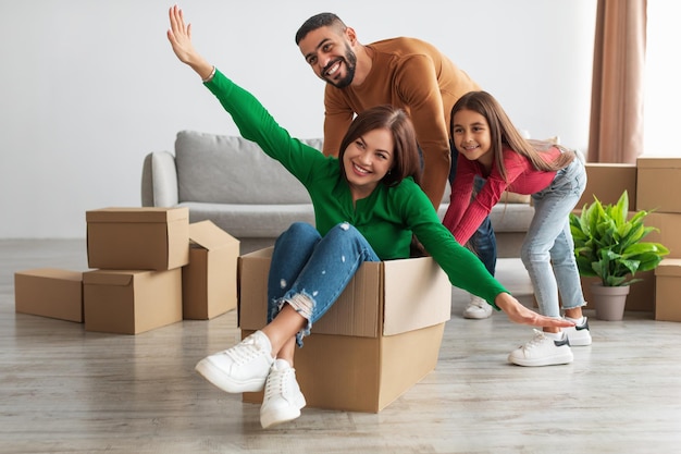 Célébrer le jour du déménagement. Portrait d'une famille arabe heureuse s'amusant ensemble dans un nouvel appartement, déballant, souriant mari chevauchant une femme excitée dans une boîte en carton dans le salon avec une petite fille