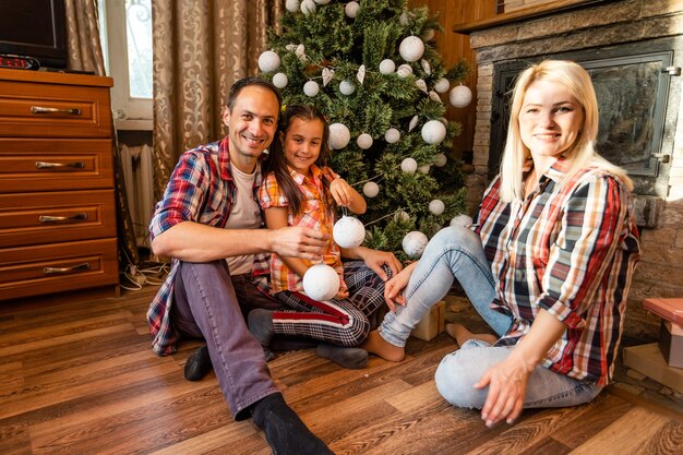 Cercle de famille à thème Noël et nouvel an. Jeune famille caucasienne assise sur un plancher en bois à la maison dans le salon près de l'arbre de Noël de la cheminée.