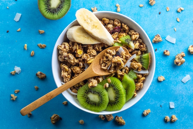 Céréales sèches pour le petit-déjeuner. Bol de muesli croustillant au miel avec des tranches de banane fraîche et du kiwi sur fond bleu. Alimentation saine, en forme et en fibres. Vue de dessus. L'heure du déjeuner